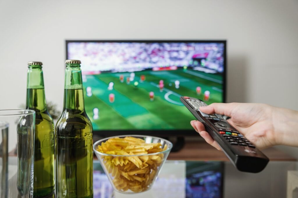 pexels-photo-1201996 A person holding a remote while watching soccer on TV with beer and snacks on the table.